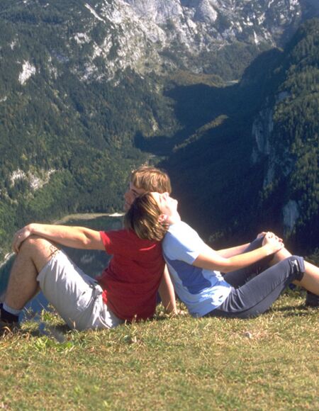 Ein Paar sitzt Rücken an Rücken in einer hoch gelegenen Wiese, mit Blick auf das Alpen-Panorama im Salzkammergut