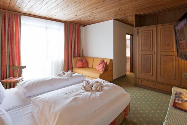 Ein Doppelbett mit weißer Bettdecke und einer kleinen Couch. Gegenüber vom Bett ist ein großer brauner Schrank