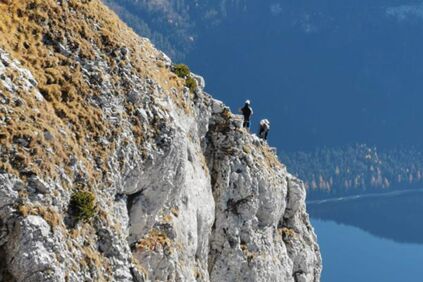 Ein Felsvorsprung auf dem zwei Menschen klettern darunter befindet sich ein See.