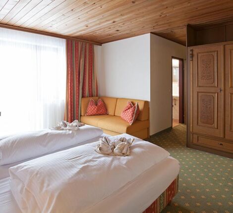 Ein Doppelbett mit weißer Bettdecke und einer kleinen Couch. Gegenüber vom Bett ist ein großer brauner Schrank