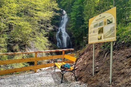 Vor einem Zaun steht eine Parkbank, dahinter steht ein Schild mit Informationen zu den "Rosas Wasserfälle".Hinter dem Zaun befinden sich die Wasserfälle.
