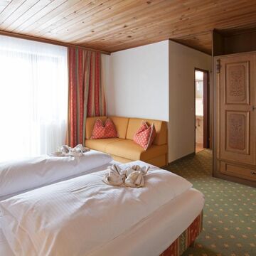 Ein Doppelbett mit weißer Bettdecke und einer kleinen Couch. Gegenüber vom Bett ist ein großer brauner Schrank
