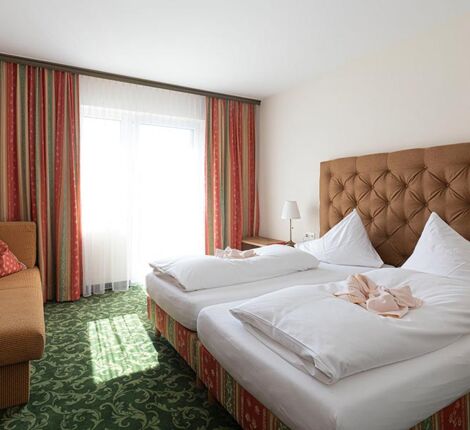Ein Hotelzimmer mit grünem Teppich und einem Doppelbett. Gegenüber ist ein Sofa und daneben ein Fenster mit Vorhängen.