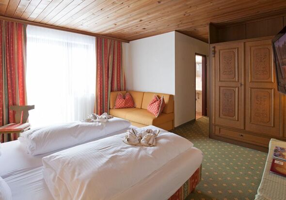 A double bed with a white duvet and a small couch. Opposite the bed is a large brown wardrobe.