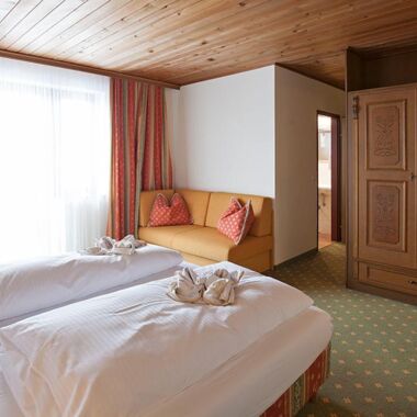 Ein Doppelbett mit weißer Bettdecke und einer kleinen Couch. Gegenüber vom Bett ist ein großer brauner Schrank