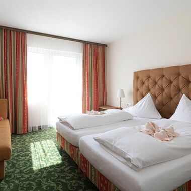 Ein Hotelzimmer mit grünem Teppich und einem Doppelbett. Gegenüber ist ein Sofa und daneben ein Fenster mit Vorhängen.