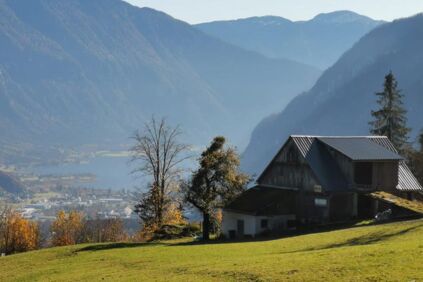 Ausblick auf einen See zwischen den Alpen im Hintergrund und einem Berghof im Vordergrund