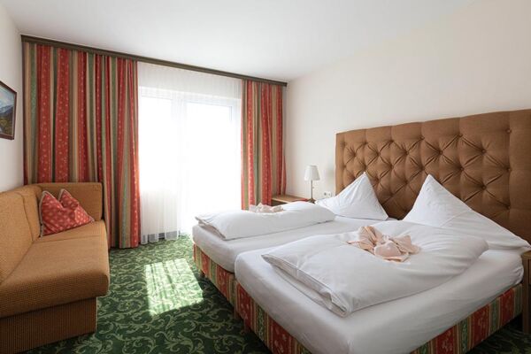 Ein Hotelzimmer mit grünem Teppich und einem Doppelbett. Gegenüber ist ein Sofa und daneben ein Fenster mit Vorhängen.