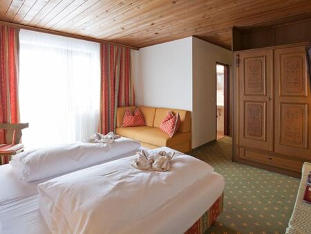 A double bed with a white duvet and a small couch. Opposite the bed is a large brown wardrobe.