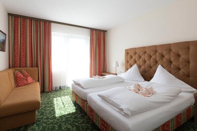 Ein Hotelzimmer mit grünem Teppich und einem Doppelbett. Gegenüber ist ein Sofa und daneben ein Fenster mit Vorhängen.