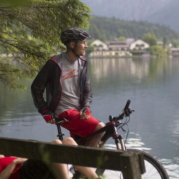 Eine Frau liegt auf der Parkbank am See und neben ihr sitzt ein Mann auf seinem Mountainbike