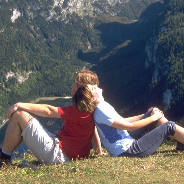 Ein Paar sitzt Rücken an Rücken in einer hoch gelegenen Wiese, mit Blick auf das Alpen-Panorama im Salzkammergut
