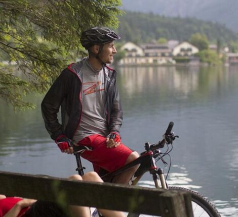 Eine Frau liegt auf der Parkbank am See und neben ihr sitzt ein Mann auf seinem Mountainbike