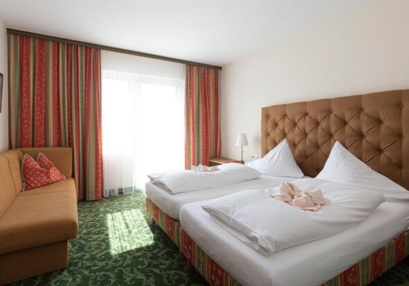 A hotel room with green carpet and a double bed. Opposite is a sofa and next to it a window with curtains.